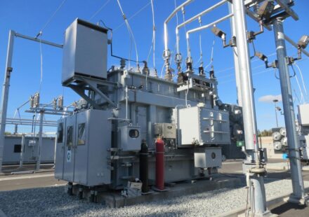 A large grey electrical transformer with metal pipes and cables in a substation