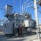 A large grey electrical transformer with metal pipes and cables in a substation