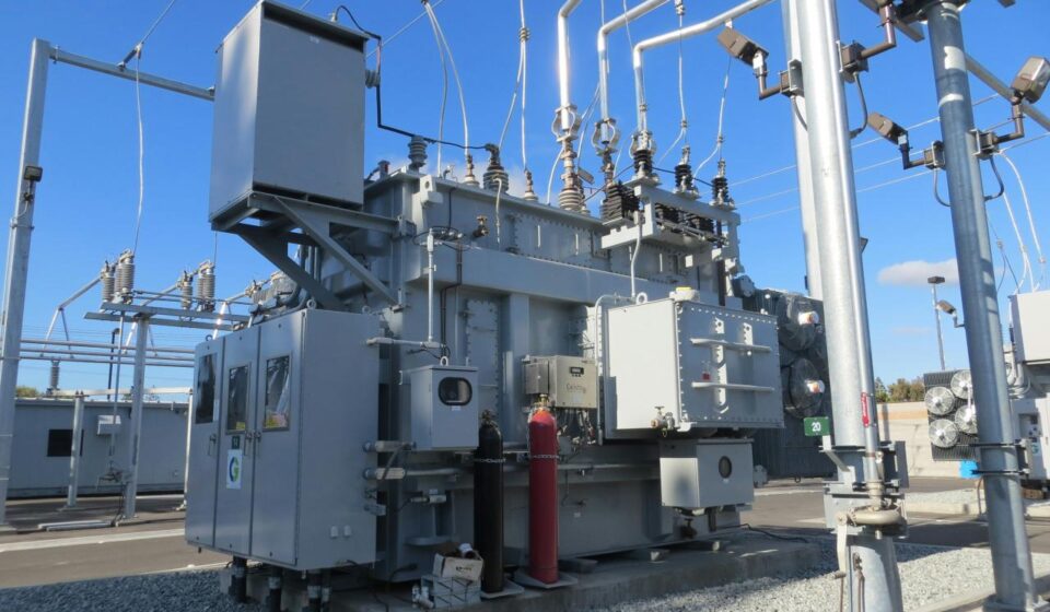 A large grey electrical transformer with metal pipes and cables in a substation